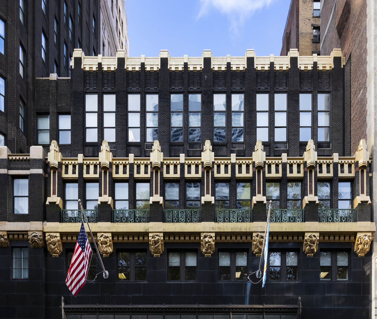 A color photograph of a lower portion of the American Radiator Building. The bottom portion is made of large black stone blocks. Above this is a continuous gold horizontal band supported by brackets with small figural sculptures. The next several stories are made of black brick and accented with a repeating rectangular pattern of golden accents.