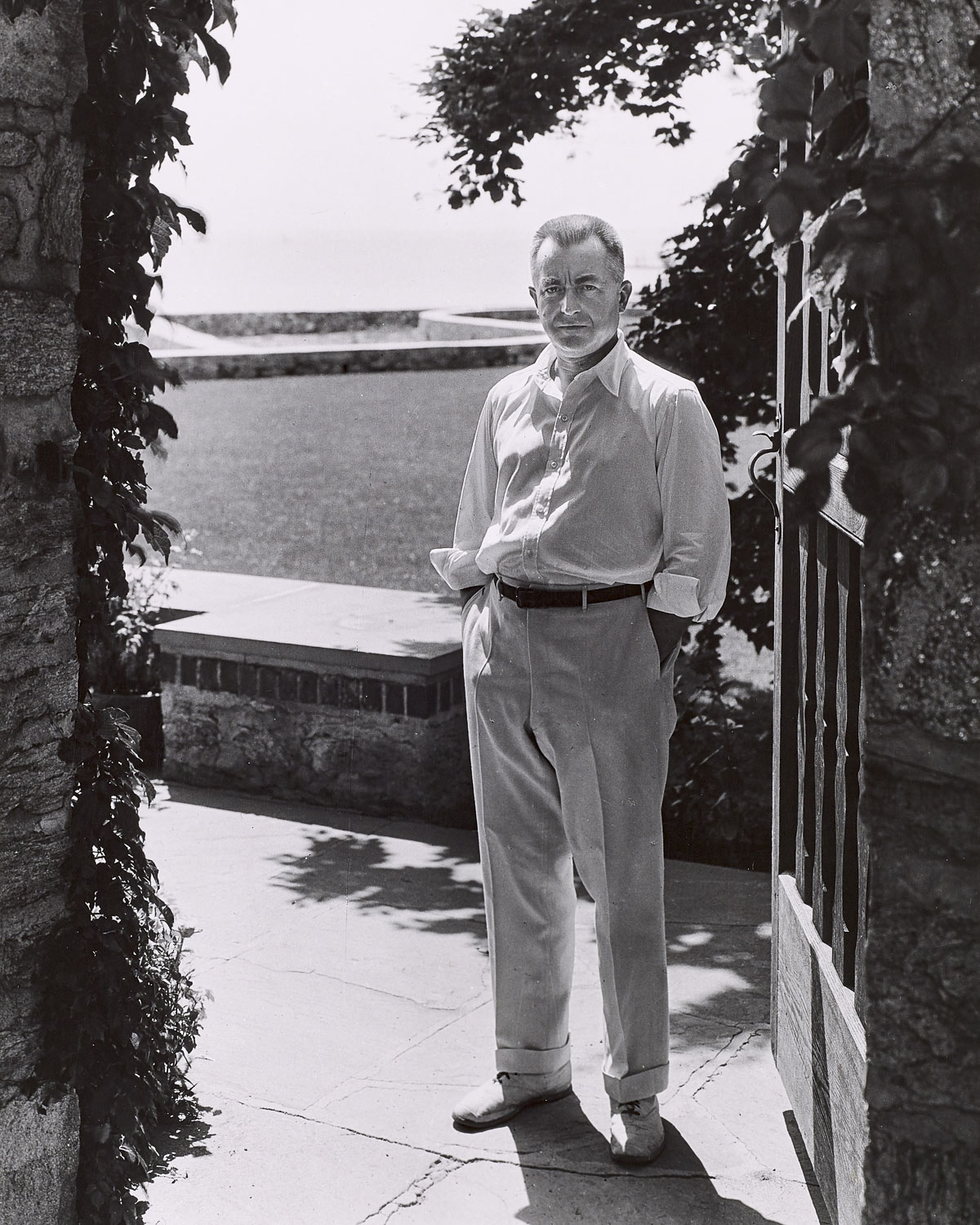 Full-length portrait of a man standing next to an open gate. Foliage is visible on the left side; there is a low stone wall directly behind him and a grassy field beyond. The man looks directly at the camera with a calm attitude, his hands in his pockets. He is wearing a white open collared shirt and light-colored cuffed pants.