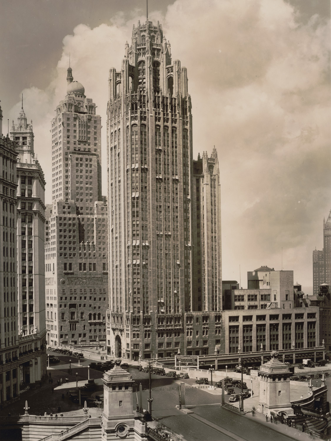 A black and white photograph of the tower as it appeared in the 1930s. Old-fashioned cars line the road that the entrance of the skyscraper is on. A bank of the Chicago River is barely visible in the lower right corner, peeking out from under a bridge. The majority of the skyscraper is the unornamented central shaft with beveled or angled corners; the base is made up of four stories topped by gothic-style balconies. The uppermost portion of the tower consists of eight flying buttresses that support a small, octagonal top portion which all together looks like a crown.