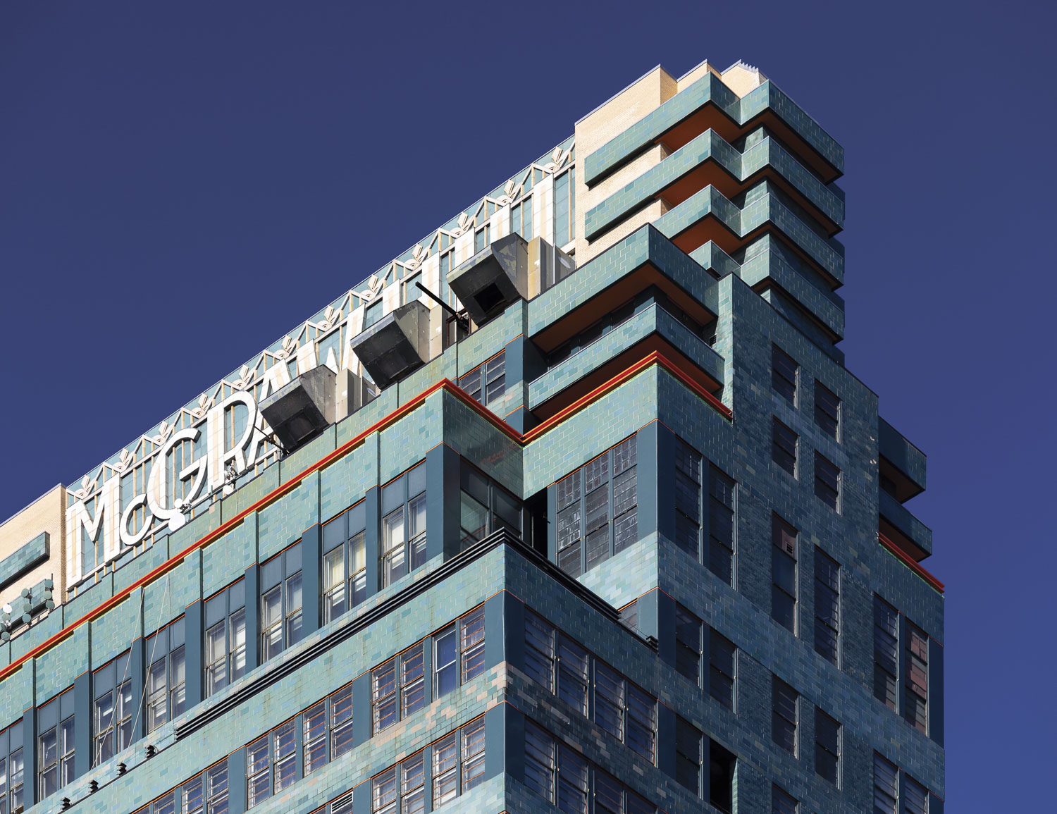 A detail photograph of the top of a skyscraper taken from a low angle. Horizontal bands of blue-gray windows alternate with continuous strips of blue-green terra cotta tiles. The company name is spelled out in large white terra cotta letters at the apex: “McGRAW-HILL.” Narrow lines of bright orange and small areas of tan adorn the facade. Boxy shapes at either end of the roof sign provide additional ornament.