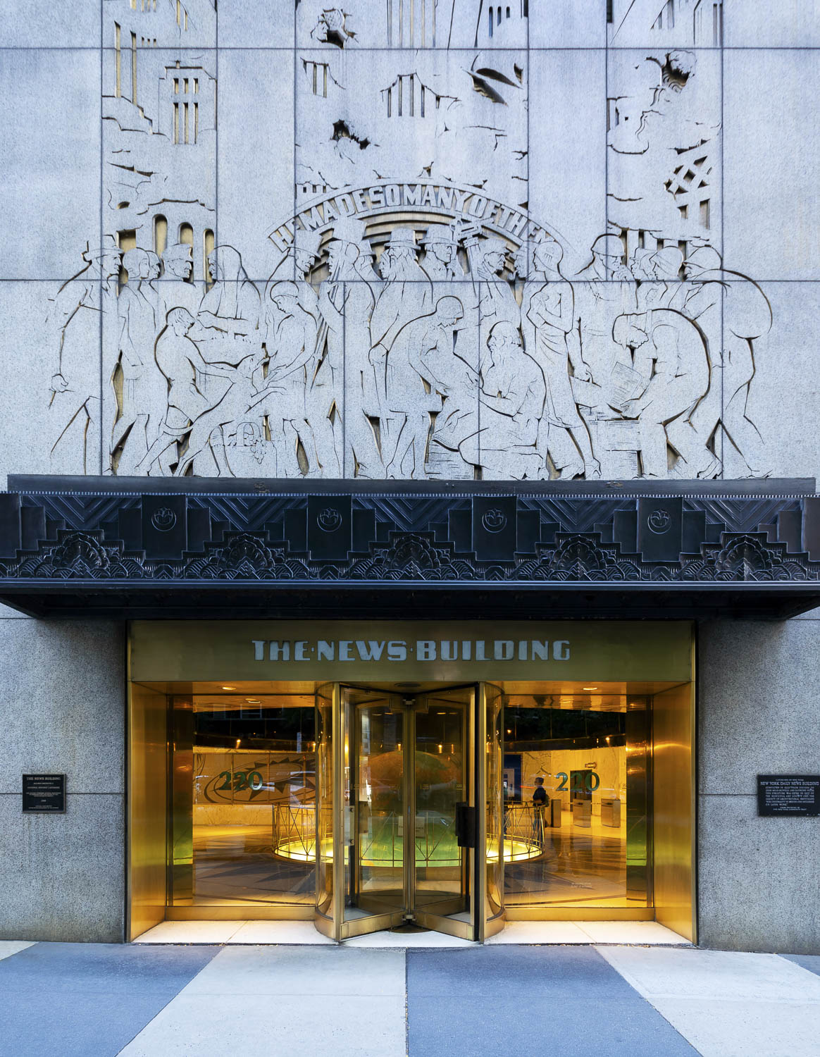 A color photograph of the entrance of a building. The lower portion features a revolving door with golden frames. Above the door is the name of the skyscraper: “The News Building.” The lobby of the building with a large globe is visible through the revolving doors. Above the entryway is a narrow metal overhand with a repeating geometric patter. Above this is a large surface of light-colored stone sculpted with the silhouettes of two dozen people of all ages dressed differently and carrying tools. The sculpture is very flat, as if each of the pieces are cutouts. A golden background shines through.