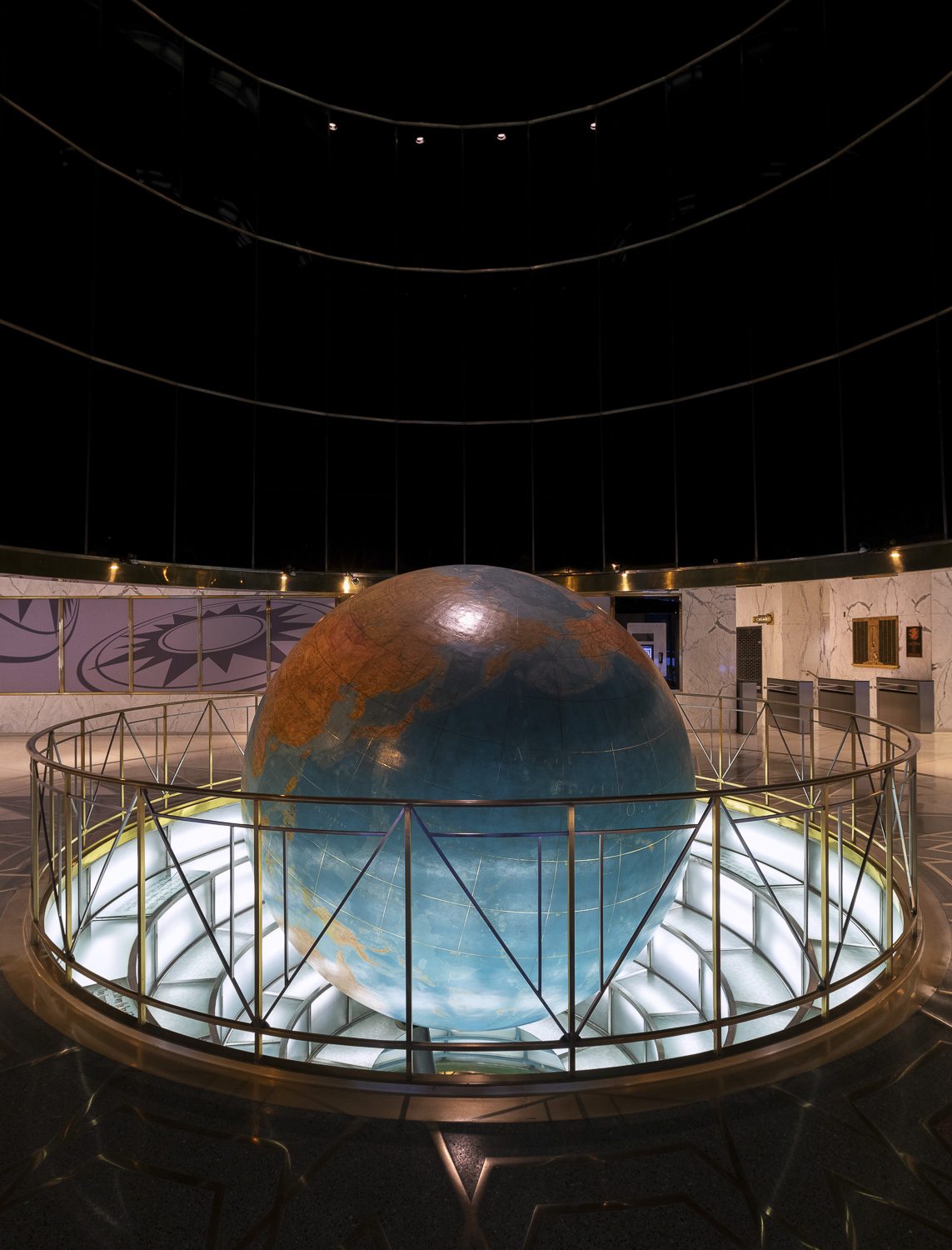 A color photograph of a portion of the lobby with a blue and green-brown globe of the earth in the center. The globe is sunken into a circular pit with stepped sides made of a translucent material that glows and illuminates the globe from below. The pit is surrounded by a simple brass railing of vertical, horizontal, and diagonal bars.