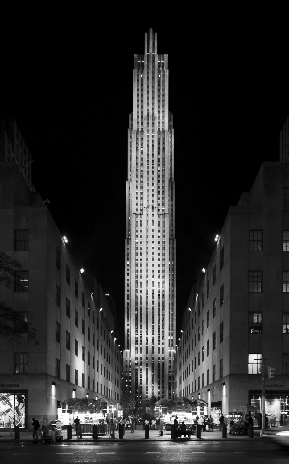 A black and white photograph of one face of a tall, narrow skyscraper at night. The tower has several small setbacks and is brilliantly illuminated, standing in contrast to the black night sky. The view of the tower is from a perpendicular main rod, looking down a broad, landscaped alley flanked by low, partly lit buildings. People hurry past storefronts on the sidewalk.