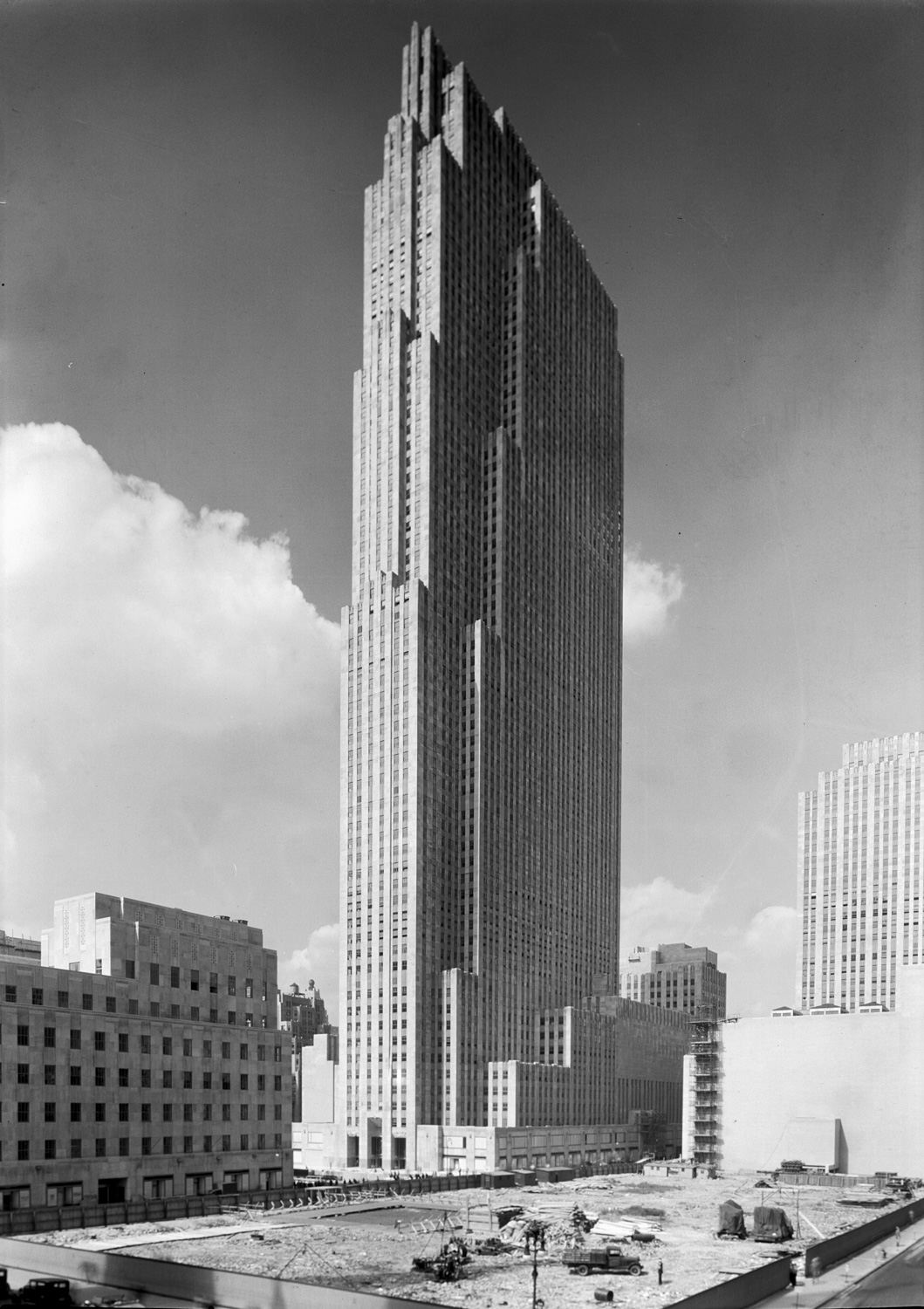 A black and white photograph of a skyscraper taken from an angle so two faces are visible. One face is very narrow and the over face is very wide. The slab-like building has several minimal setbacks and is mostly unornamented. It is made of a light-colored stone. A large plot of land between the camera and the skyscraper has been cleared. On it are some workers and a truck ready to begin additional construction.