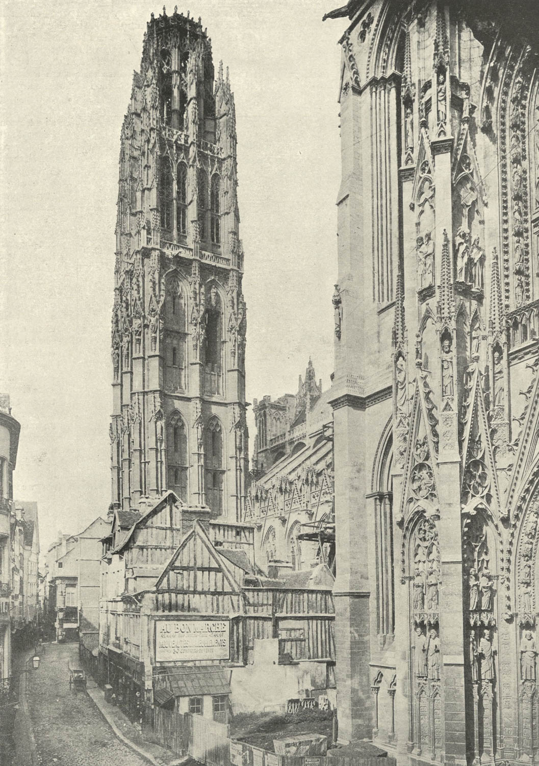 The focus of the black and white photograph is a tall stone cathedral tower set against a clear sky. The tower is roughly square, with four faces. It is covered in sculpture, tracery, and pointed arches. The uppermost portion of the tower is small and octagonal and supported by flying buttresses. In the foreground are low, timber-frame houses and shops built against the wall of the cathedral.