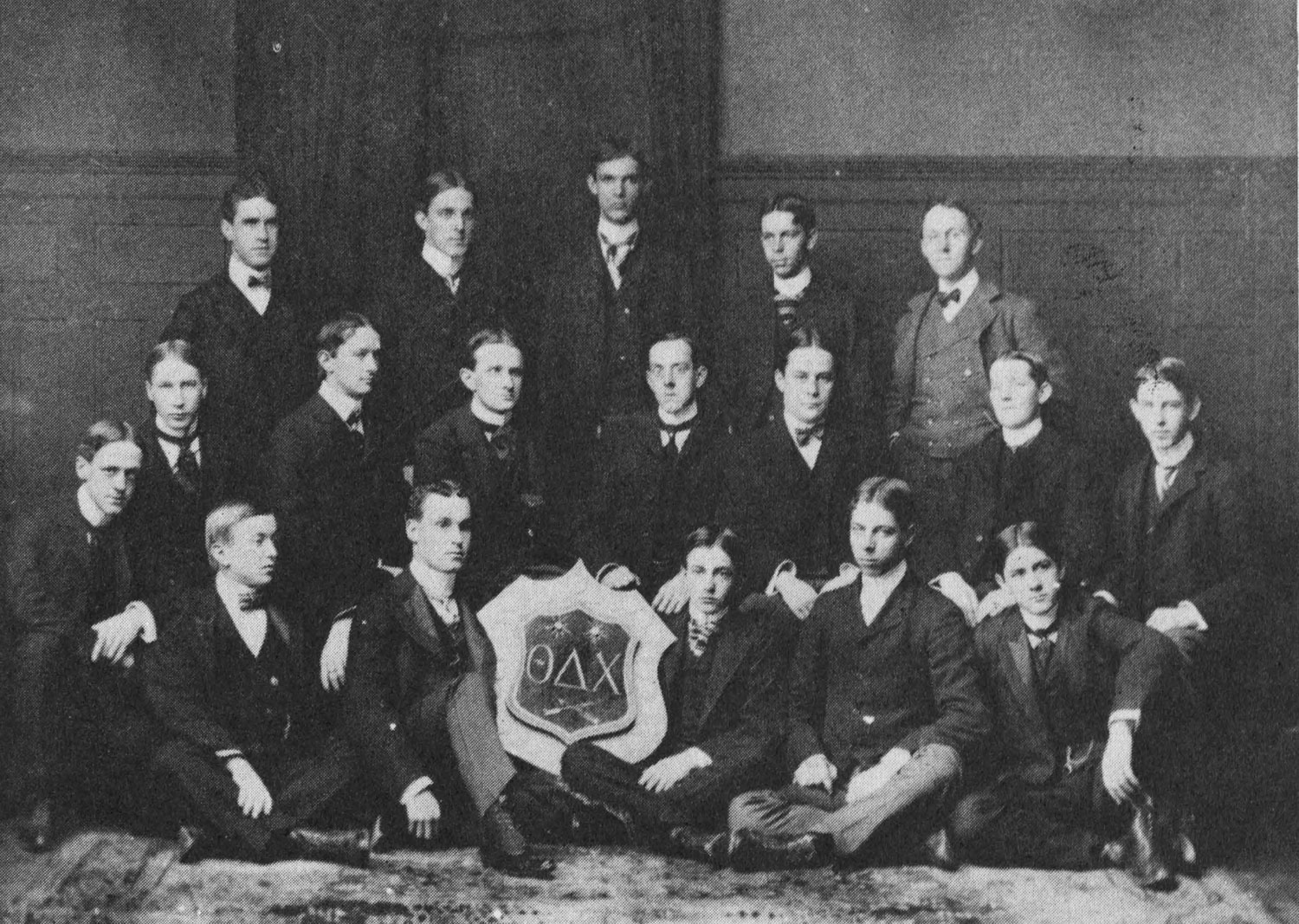 A photograph of seventeen young men standing and seated around a heraldic shield 
                    containing the Greek letters Theta, Delta, and Chi. They are dressed in dark 
                    jackets with ties. An older man in a lighter colored suit stands in the upper right.
