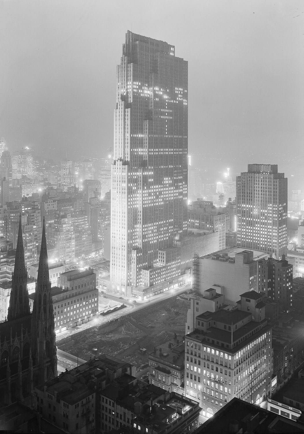A black and white photograph taken at night from a high vantage point diagonally across from Rockefeller Center. The main central skyscraper (the RCA Building) is surrounded by the lower buildings of the project. The buildings are illuminated by floodlights from the exterior and a random assortment of the windows glow with light from the interior. The windows of other city buildings twinkle in the background and disappear into a gray haze. In the lower left foreground are the darkly silhouetted spires of Saint Patrick’s Cathedral.