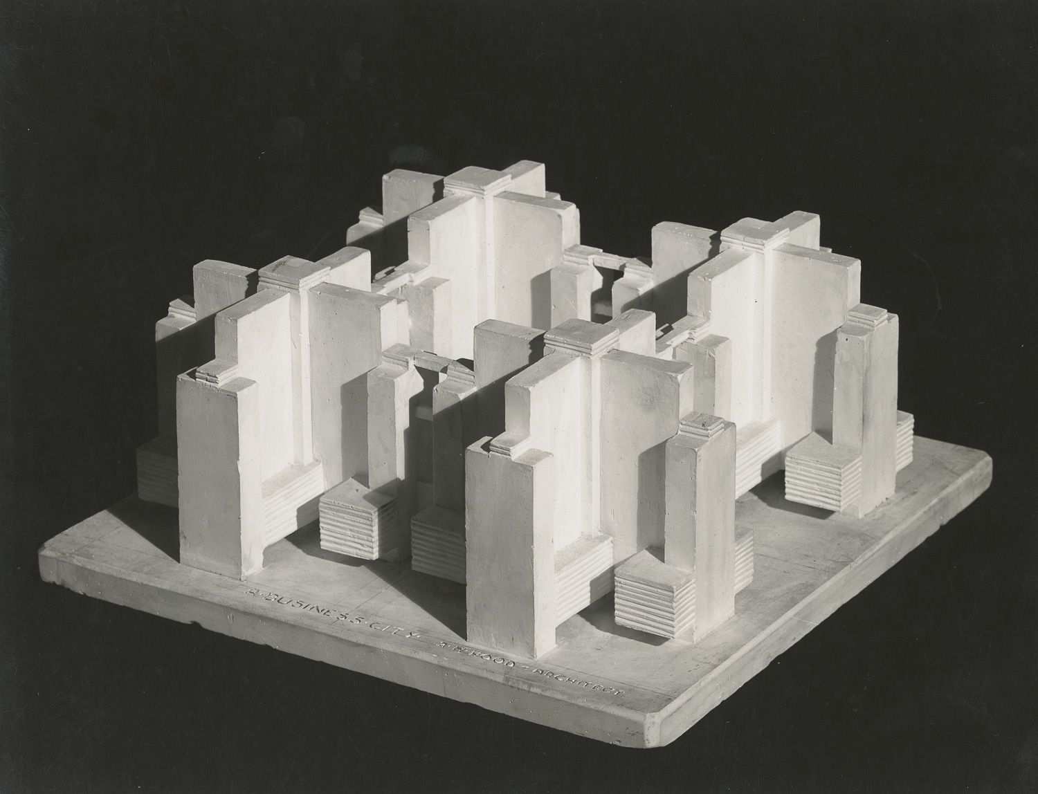A black and white photograph taken from an angle above an architectural model on a square base. The model consists of four identical towers arranged in a two-by-two grid. Each tower has a cruciform (cross-shaped) plan, with four equal wings radiating out from a central core.