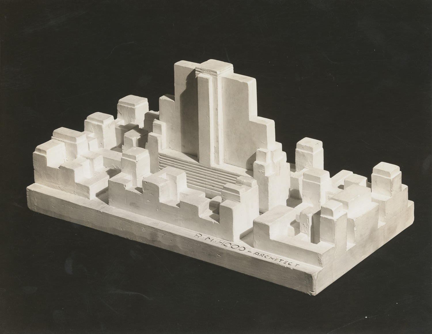 A black and white photograph taken from an angle above an architectural model on a rectangular base. At the center of a model is a building that fills an entire city block. A flattened slab with setbacks rises up from a base that fills the entire block. Surrounding this main central building are other city blocks with lower, nondescript buildings.
